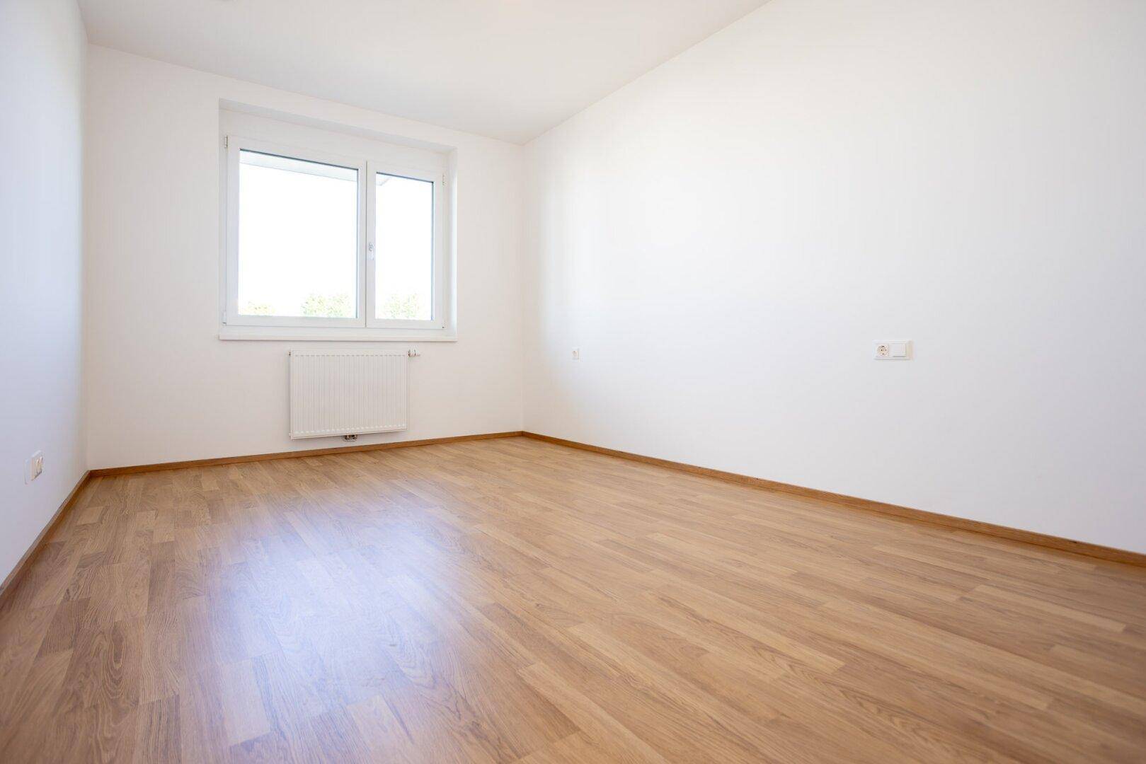 Freundliches Schlafzimmer mit Laminatboden, weißen Wänden und einem großen Fenster für viel Tageslicht.