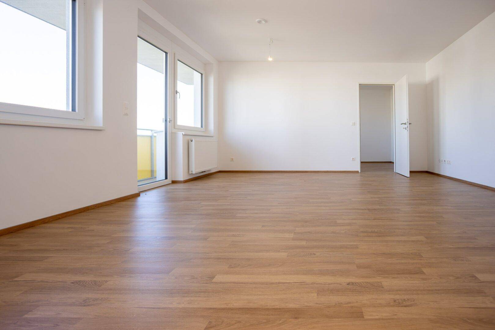 Heller Wohnbereich mit Laminatboden, weißen Wänden und direktem Zugang zum Balkon durch eine Glastür.