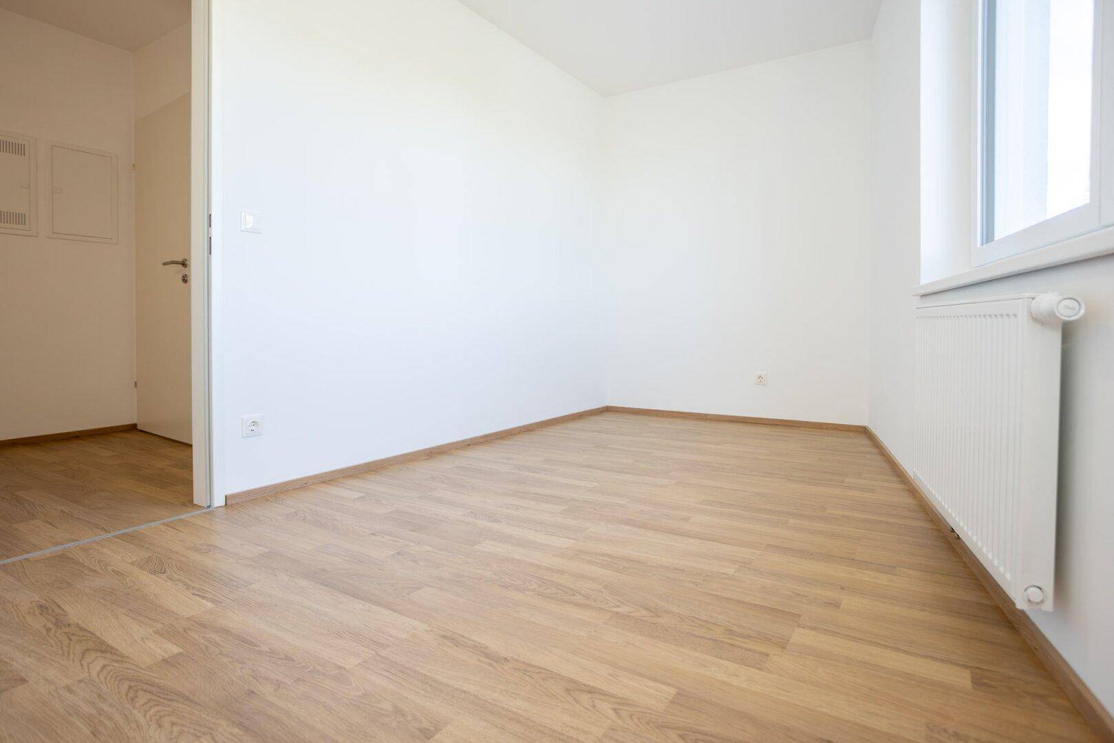 Helles Zimmer mit Laminatboden, weißen Wänden und einem Heizkörper unter dem Fenster, bereit zum Einzug.