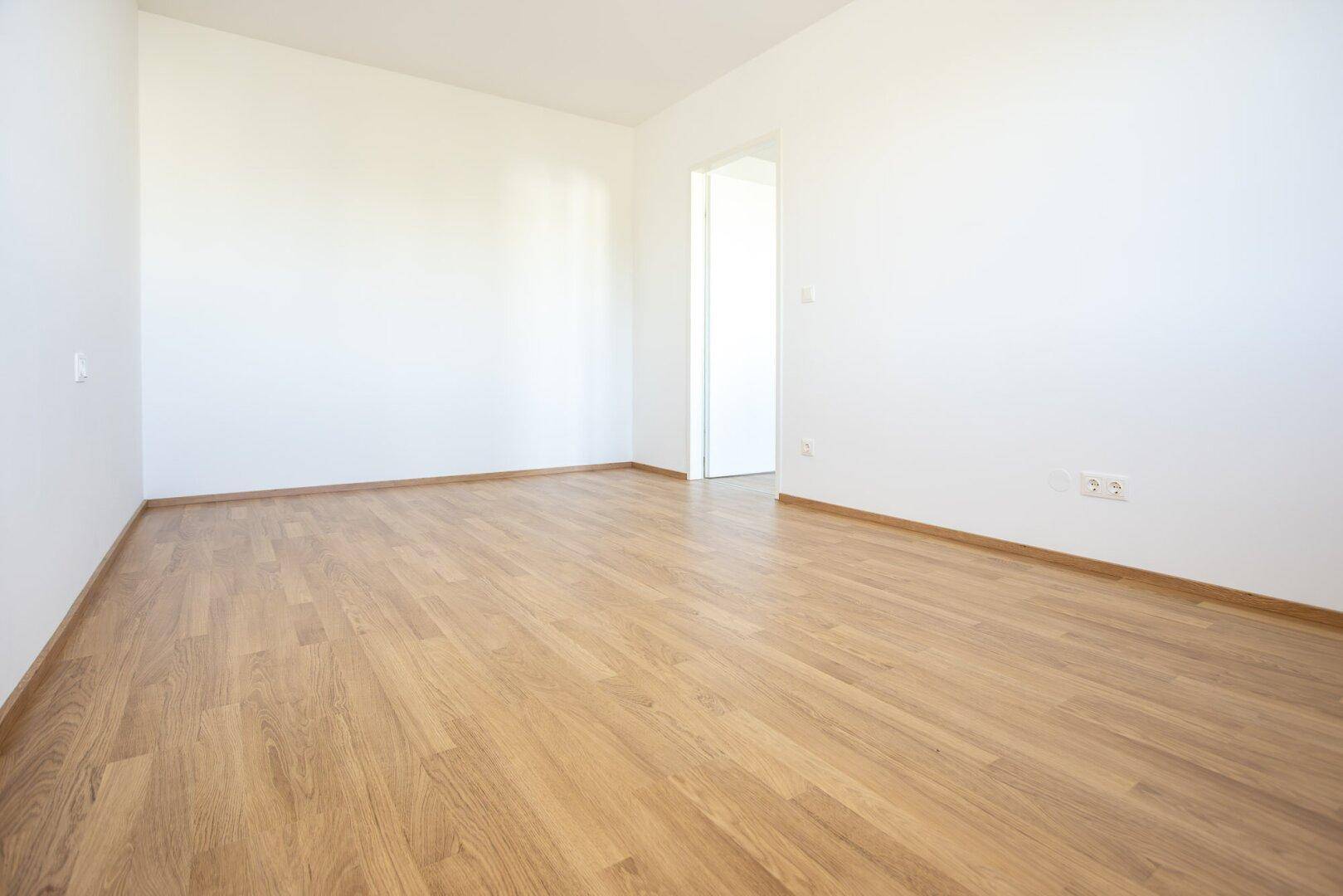 Geräumiger, leerer Raum mit hellem Laminatboden und weißen Wänden, ideal für flexible Einrichtung.