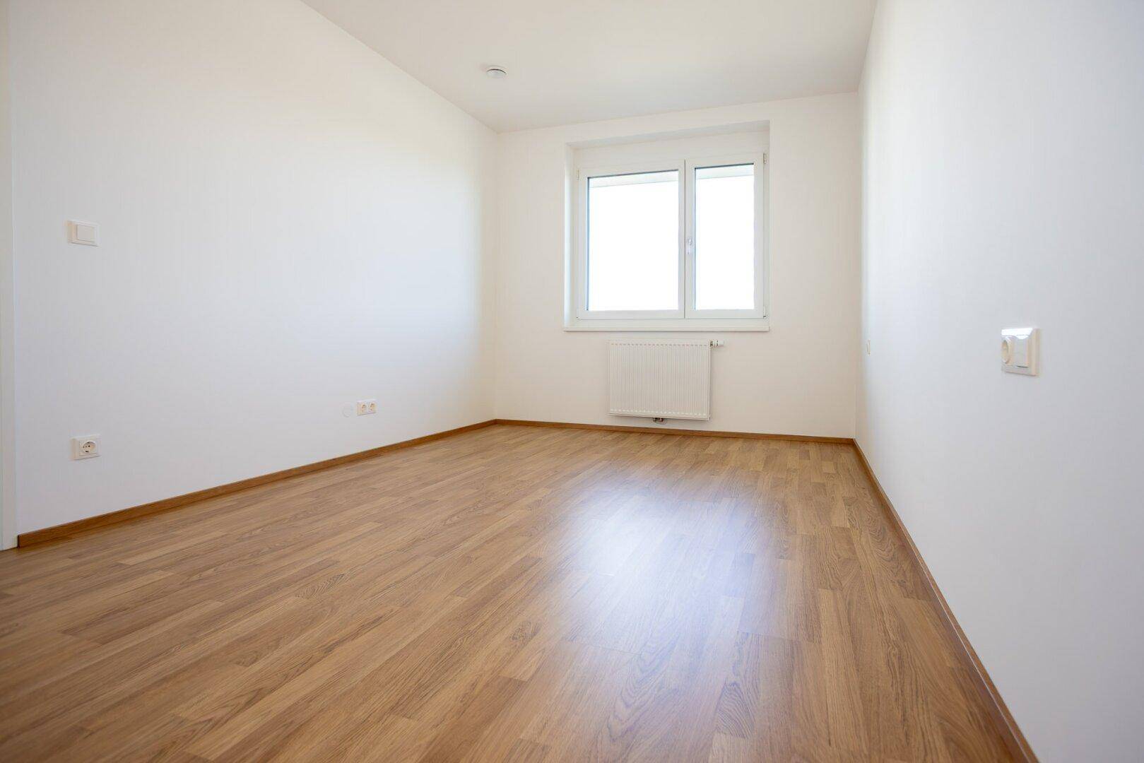Helles Zimmer mit Laminatboden, weißen Wänden und einem Fenster, das viel natürliches Licht hereinlässt.