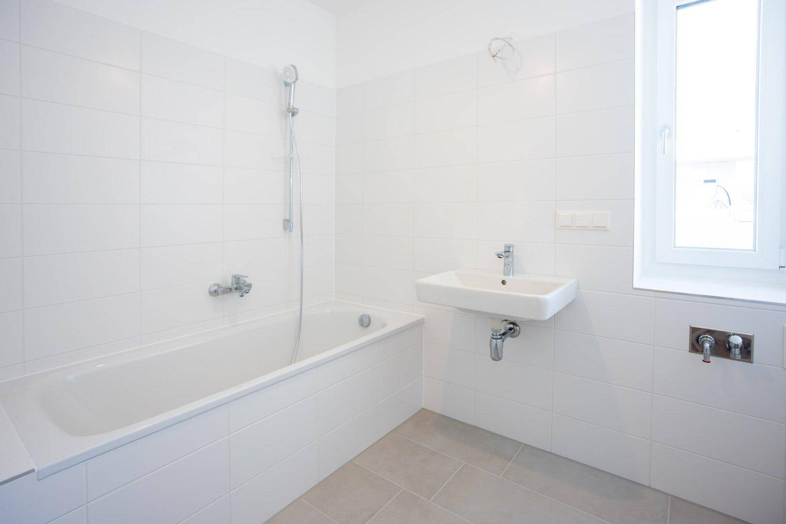 Modernes Badezimmer mit weißen Fliesen, Badewanne, Einzelwaschbecken und einem Fenster für Tageslicht.