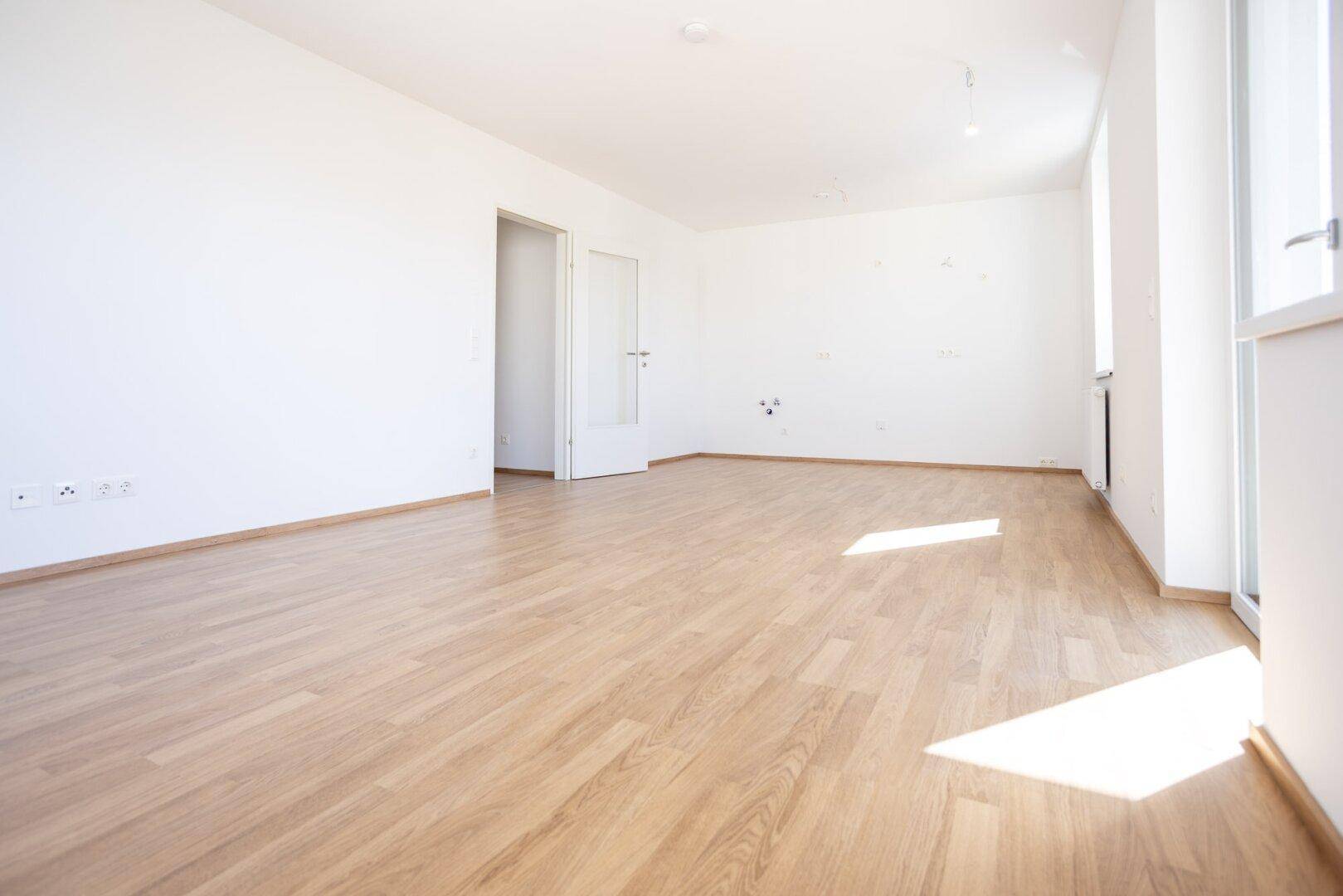 Geräumiges, helles Zimmer mit Laminatboden, weißen Wänden und viel Tageslicht durch große Fenster.