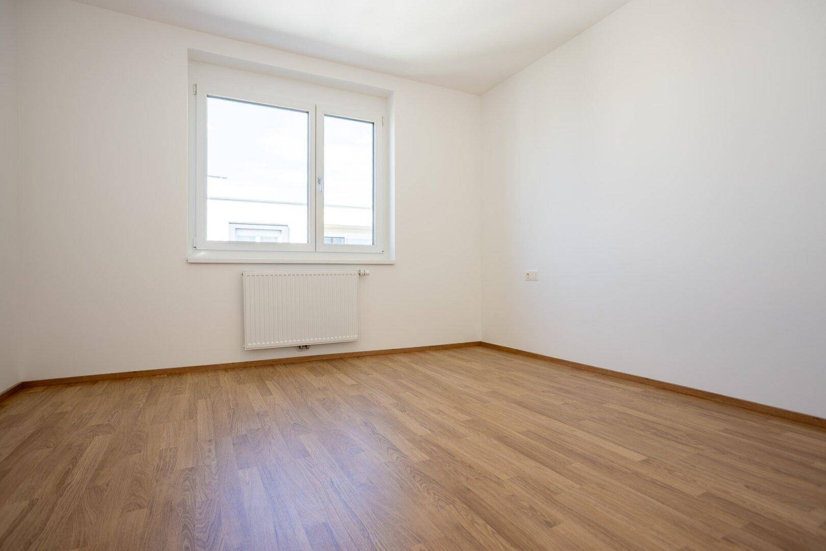 Ruhiges, helles Zimmer mit Laminatboden, weißen Wänden und einem großen Fenster.