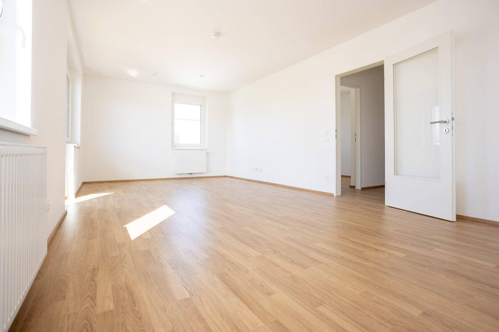 Heller, unmöblierter Raum mit Laminatboden, weißen Wänden und offener Tür zu einem weiteren Zimmer.