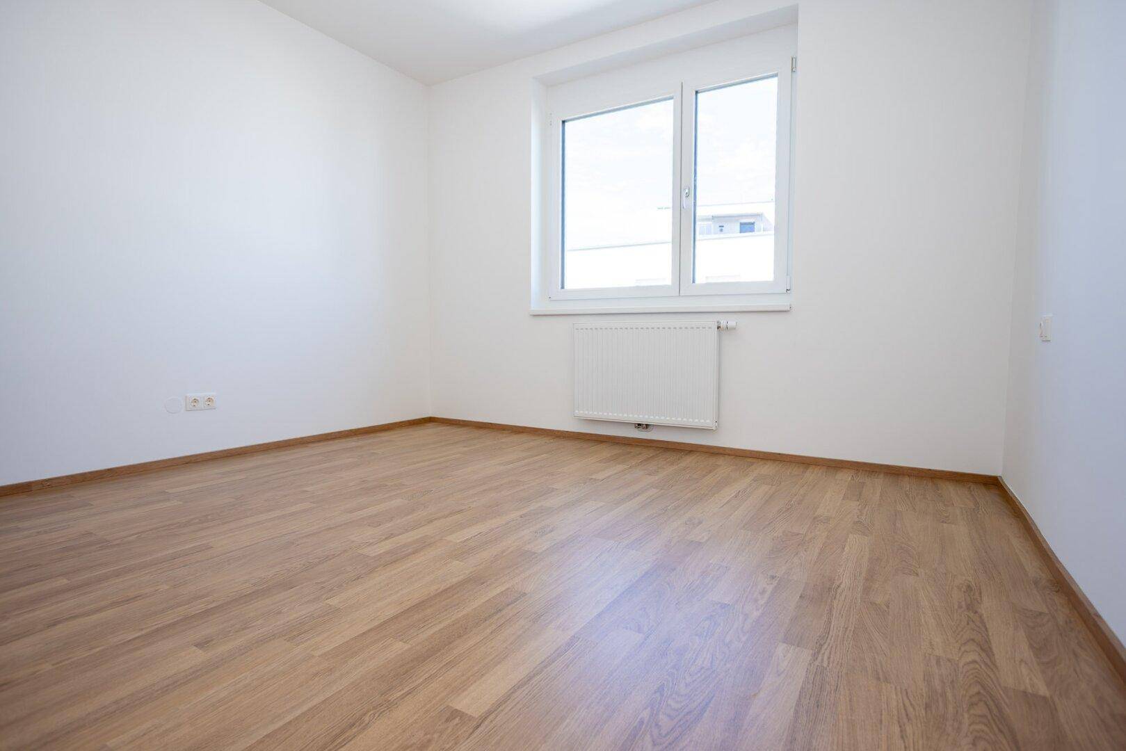 Unmöbliertes Zimmer mit hellem Laminatboden, weißen Wänden und einem Fenster für Tageslicht.