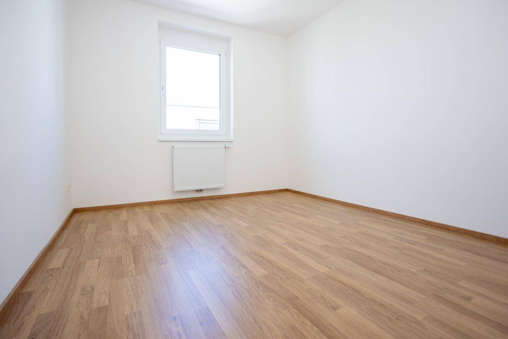 Schlichtes, helles Zimmer mit Laminatboden, weißen Wänden und einem Fenster mit Heizkörper darunter.