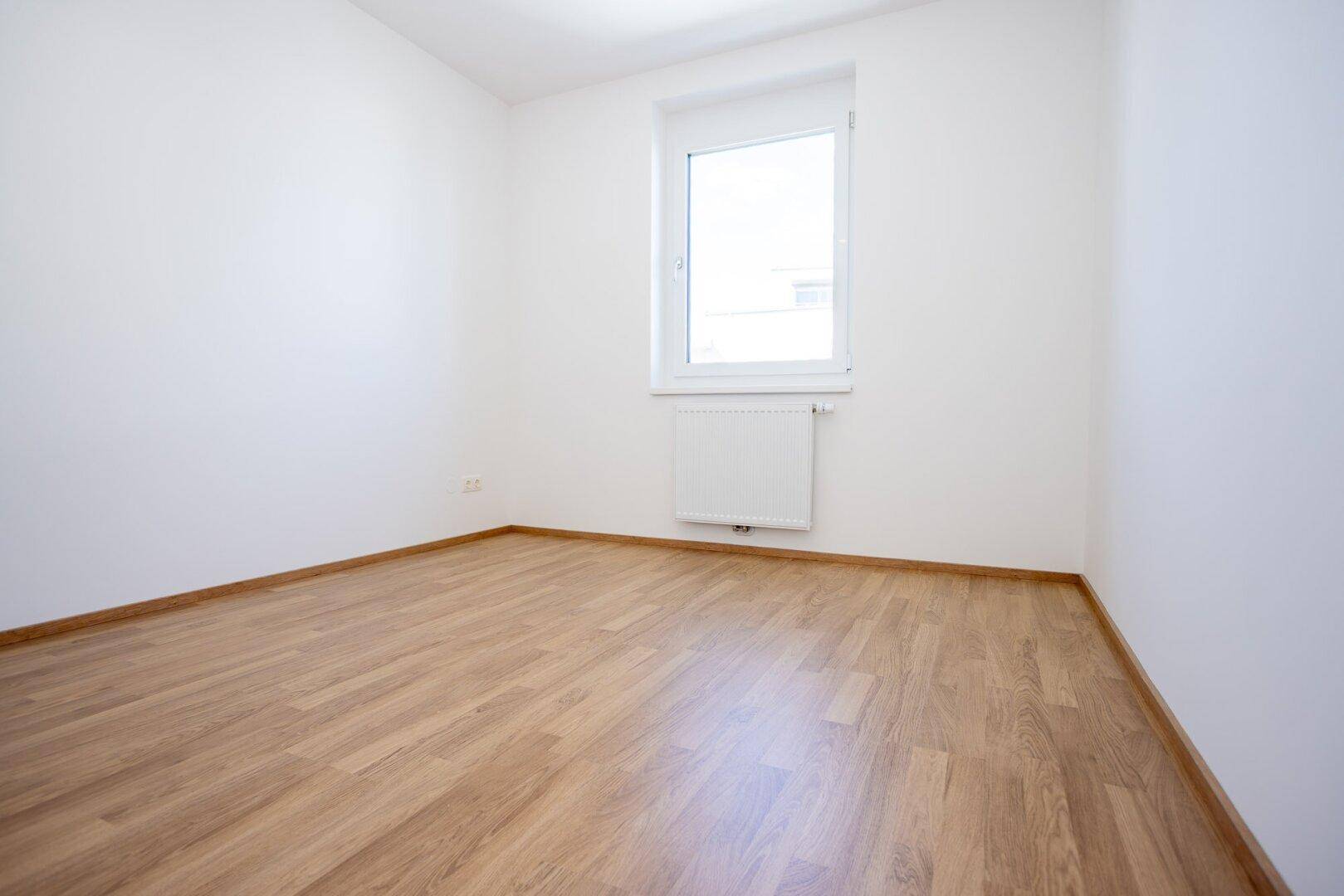 Leeres Zimmer mit Laminatboden, weißen Wänden und einem Fenster, ideal als Schlaf- oder Arbeitszimmer.