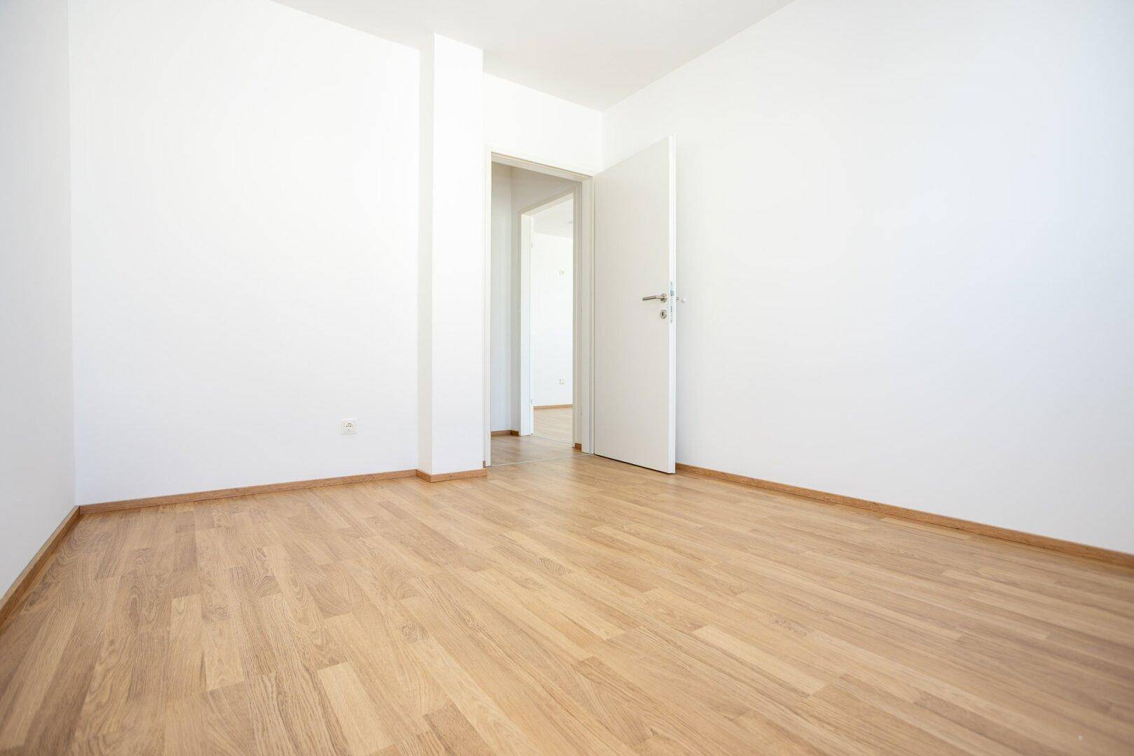 Heller, leerer Raum mit Laminatboden und weißen Wänden, der zu einem weiteren Zimmer führt.