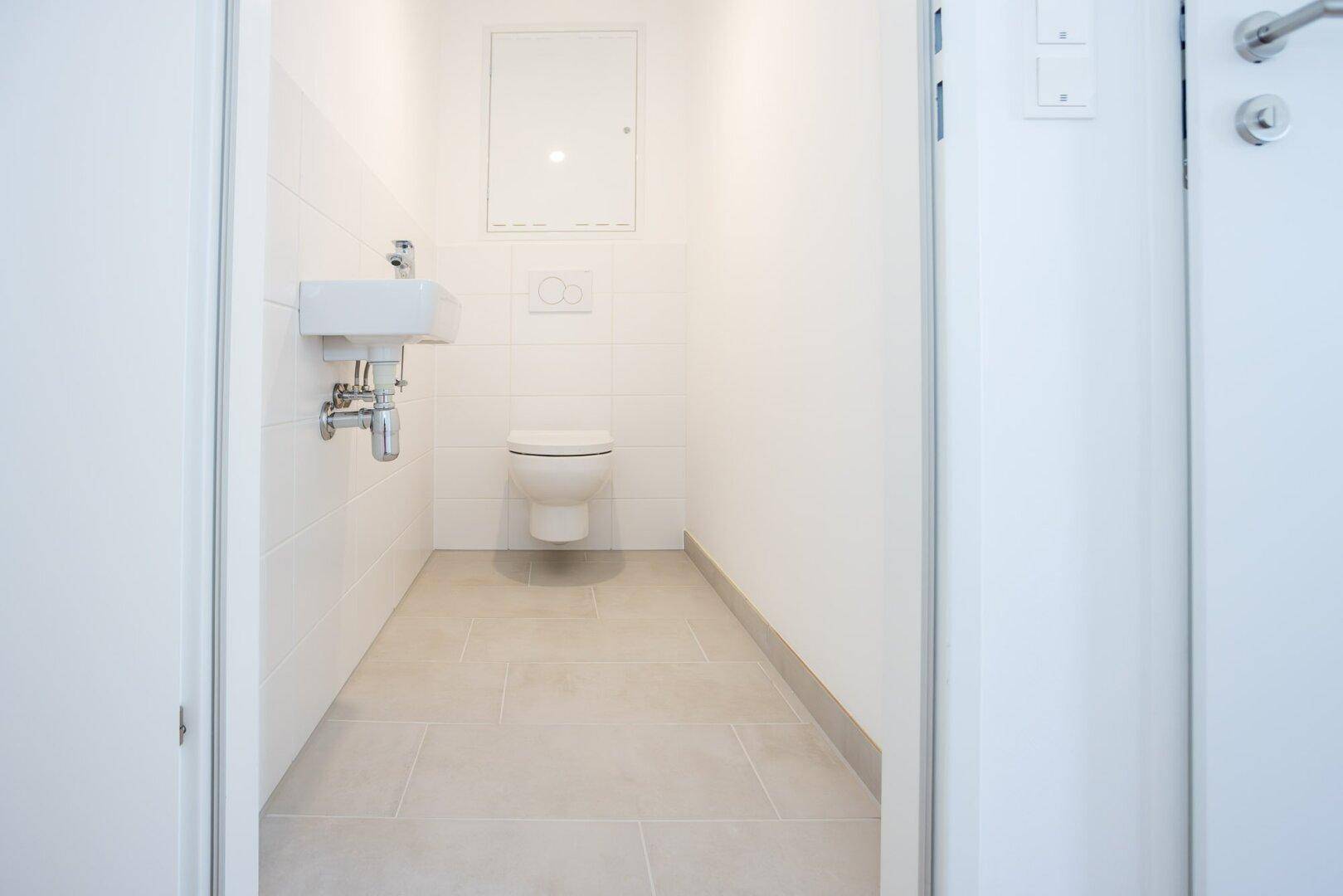 Helles Gäste-WC mit wandhängender Toilette, kleinem Waschbecken und hellen Fliesen.