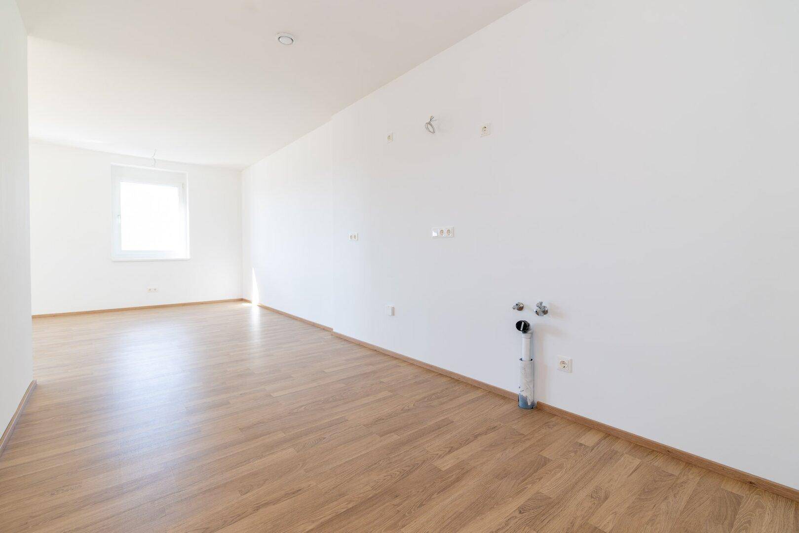 Leerer Küchenraum mit Laminatboden, weißen Wänden und vorbereiteten Anschlüssen für eine Einbauküche.
