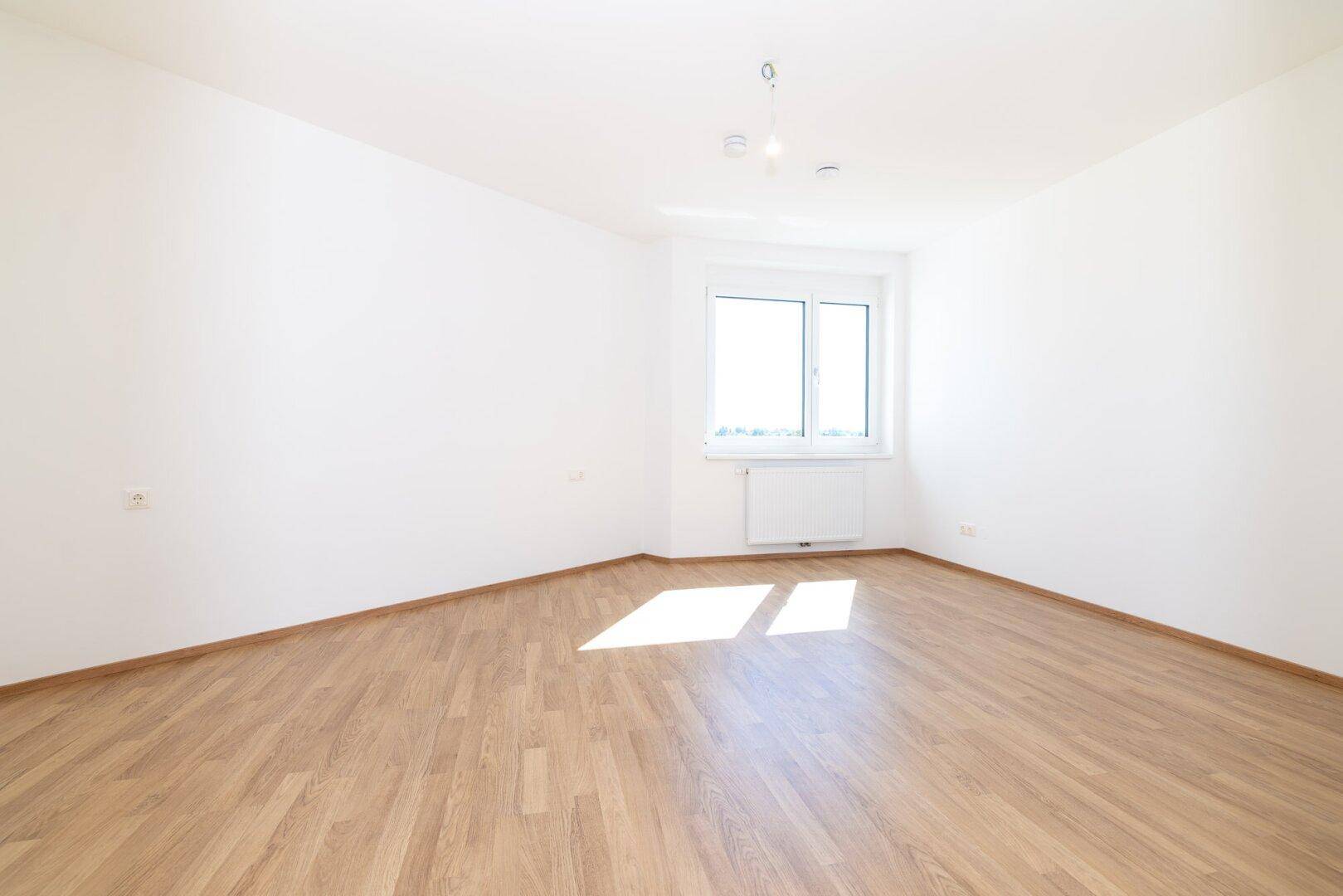 Helles Schlafzimmer mit Laminatboden, weißen Wänden und einem Fenster, das viel Tageslicht hereinlässt.