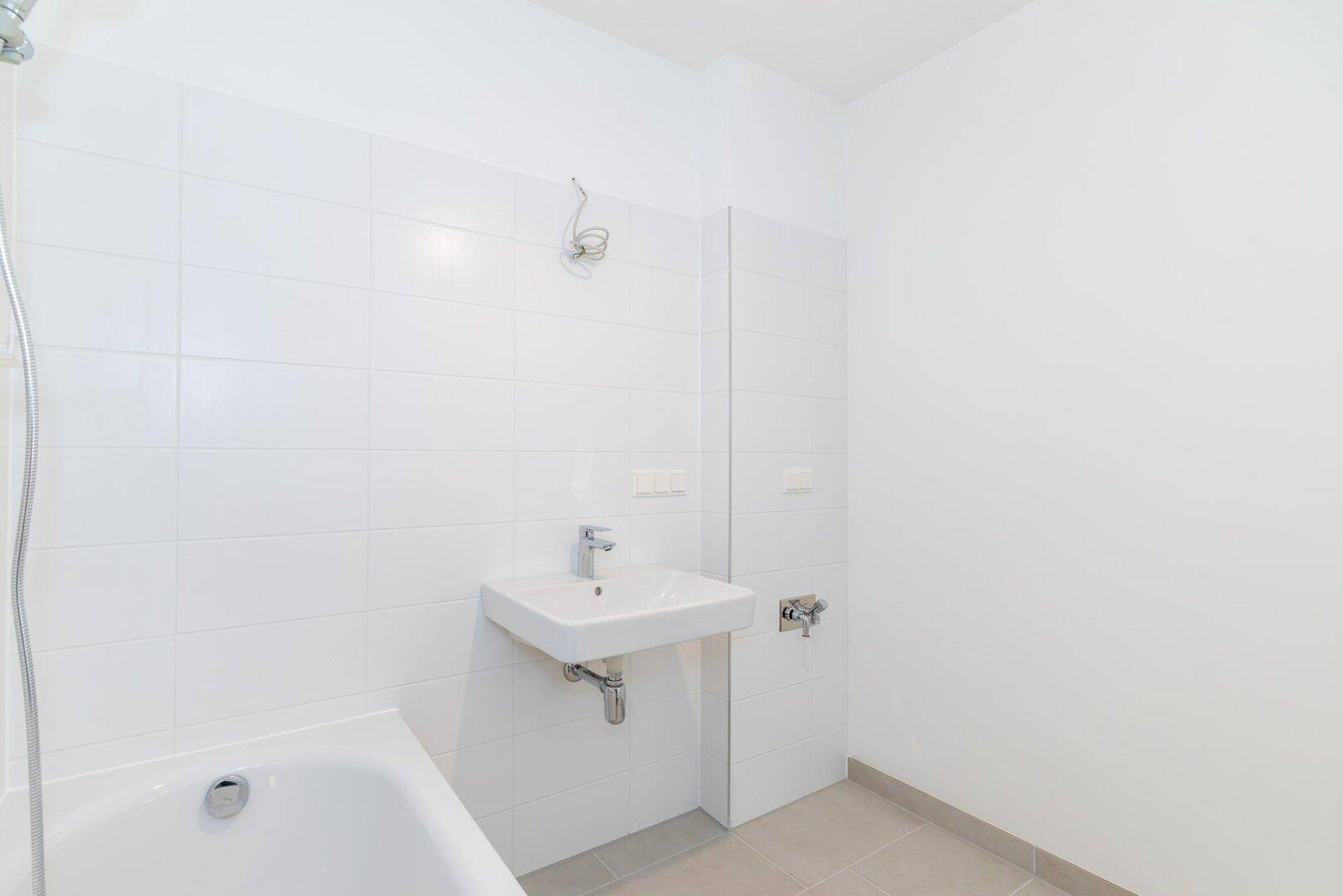 Helles Badezimmer mit weißen Fliesen, grauen Bodenfliesen, Badewanne und modernem Waschbecken.