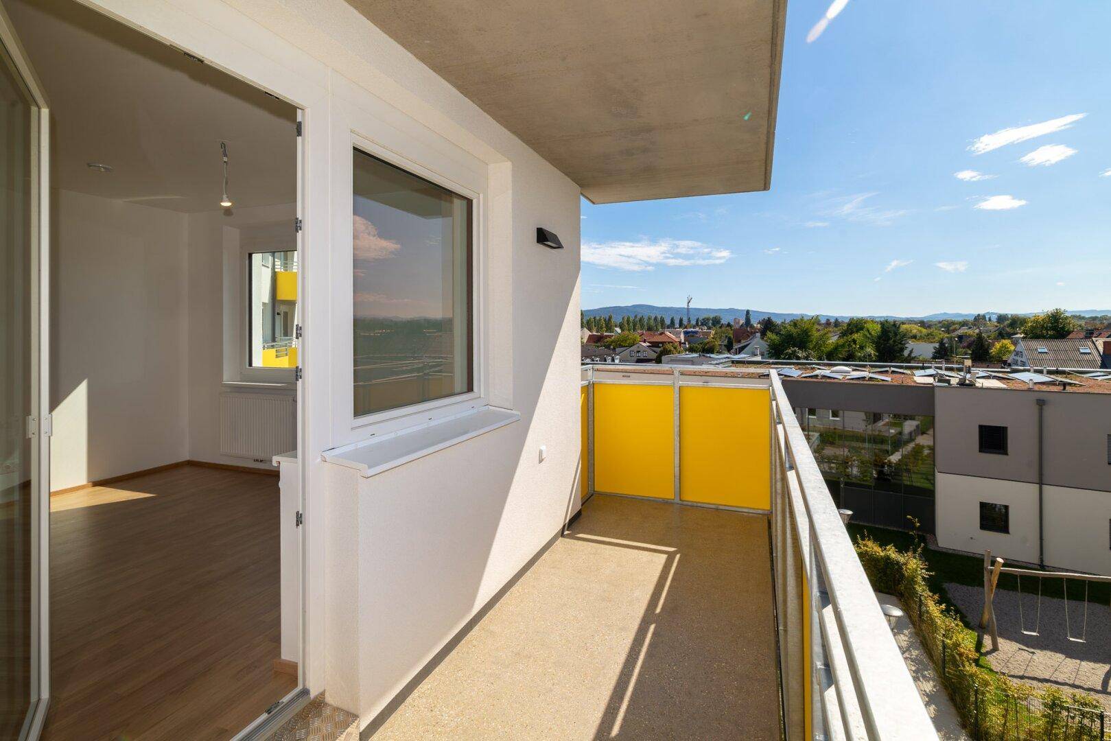 Balkon mit gelben Elementen, Blick in den Innenraum und auf eine Grünfläche mit Spielplatz.