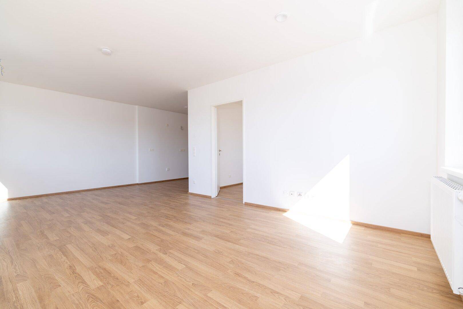 Heller, leerer Wohnraum mit Laminatboden und weißen Wänden, durchflutet von natürlichem Sonnenlicht.