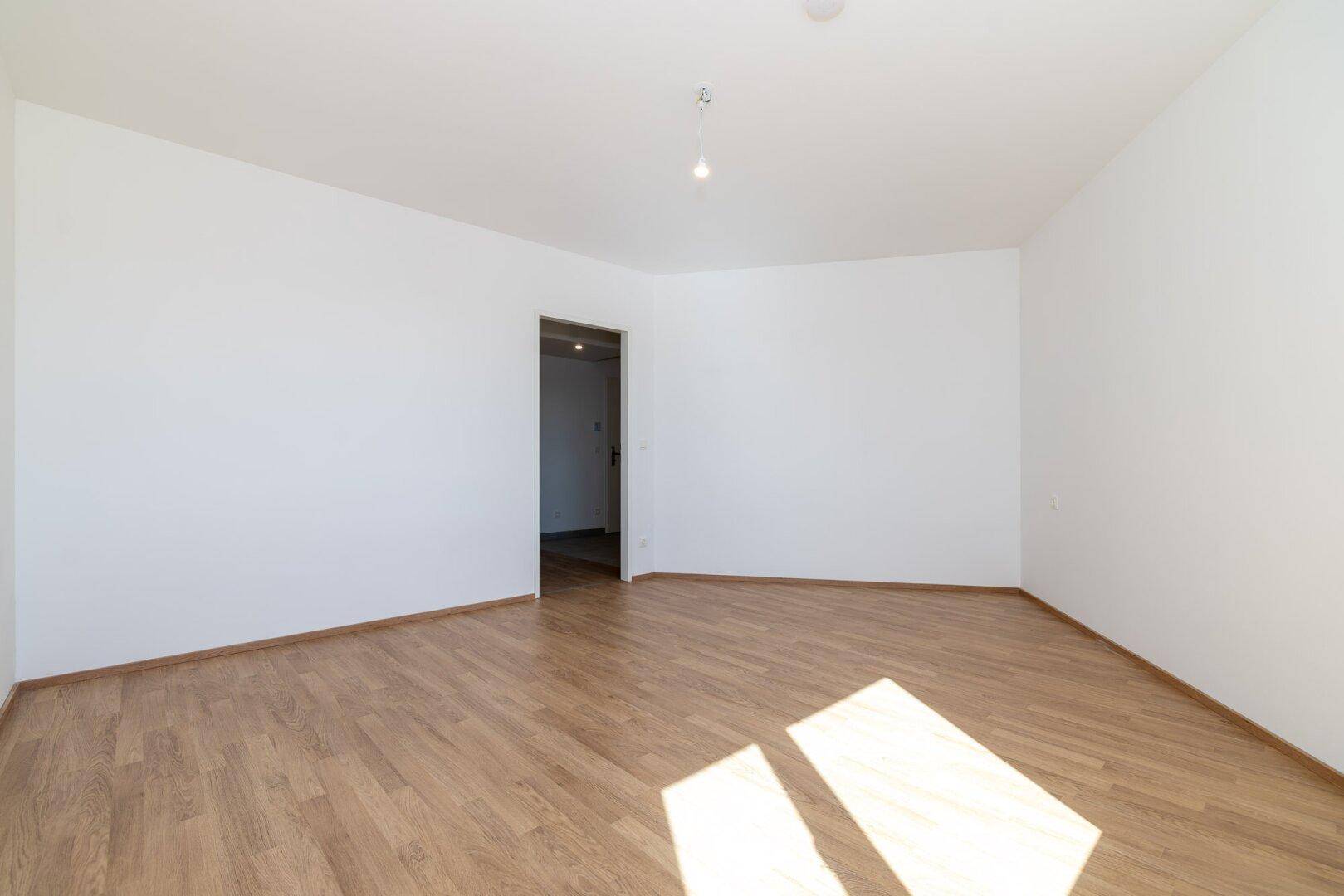 Leeres Schlafzimmer mit Laminatboden und weißen Wänden, durchflutet von natürlichem Sonnenlicht.