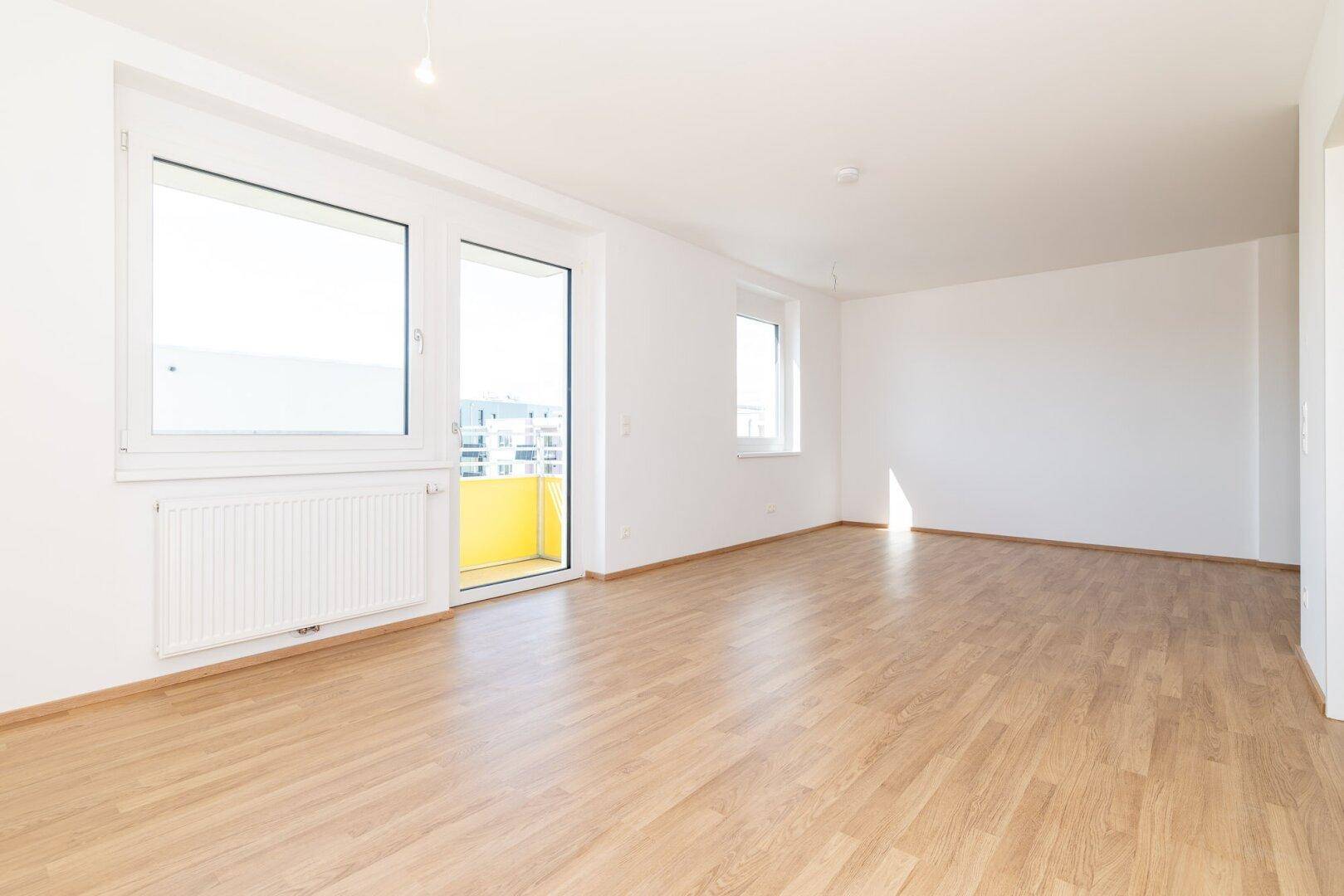 Lichtdurchfluteter Raum mit hellem Laminatboden, weißen Wänden und großem Fenster mit Balkontür.