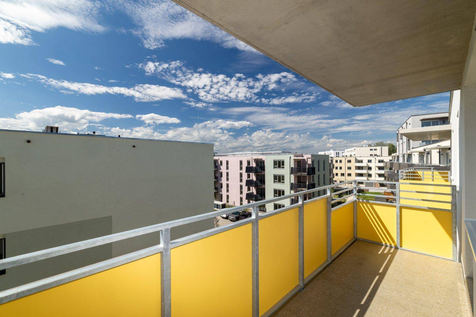 Geräumiger Balkon mit gelben Paneelen und Blick auf die moderne Wohnbebauung unter blauem Himmel.