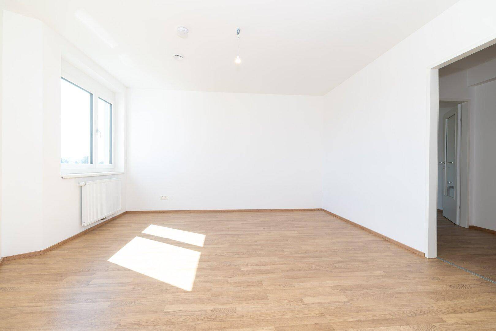 Helles Schlafzimmer mit Laminatboden, weißen Wänden und einem Fenster, das viel Tageslicht hereinlässt.
