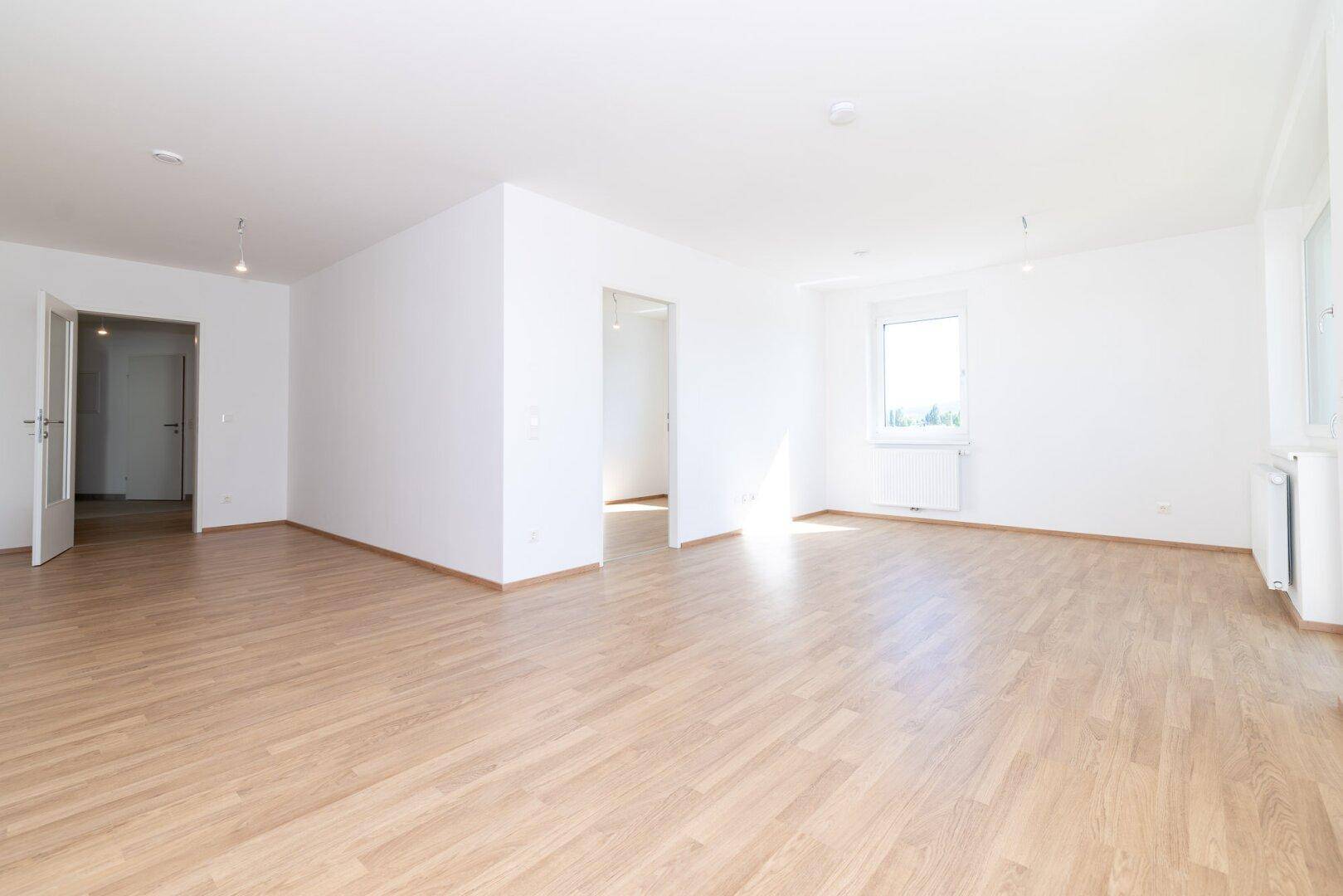 Geräumiger, heller Wohnbereich mit Laminatboden und weißen Wänden, der zu weiteren Räumen führt.