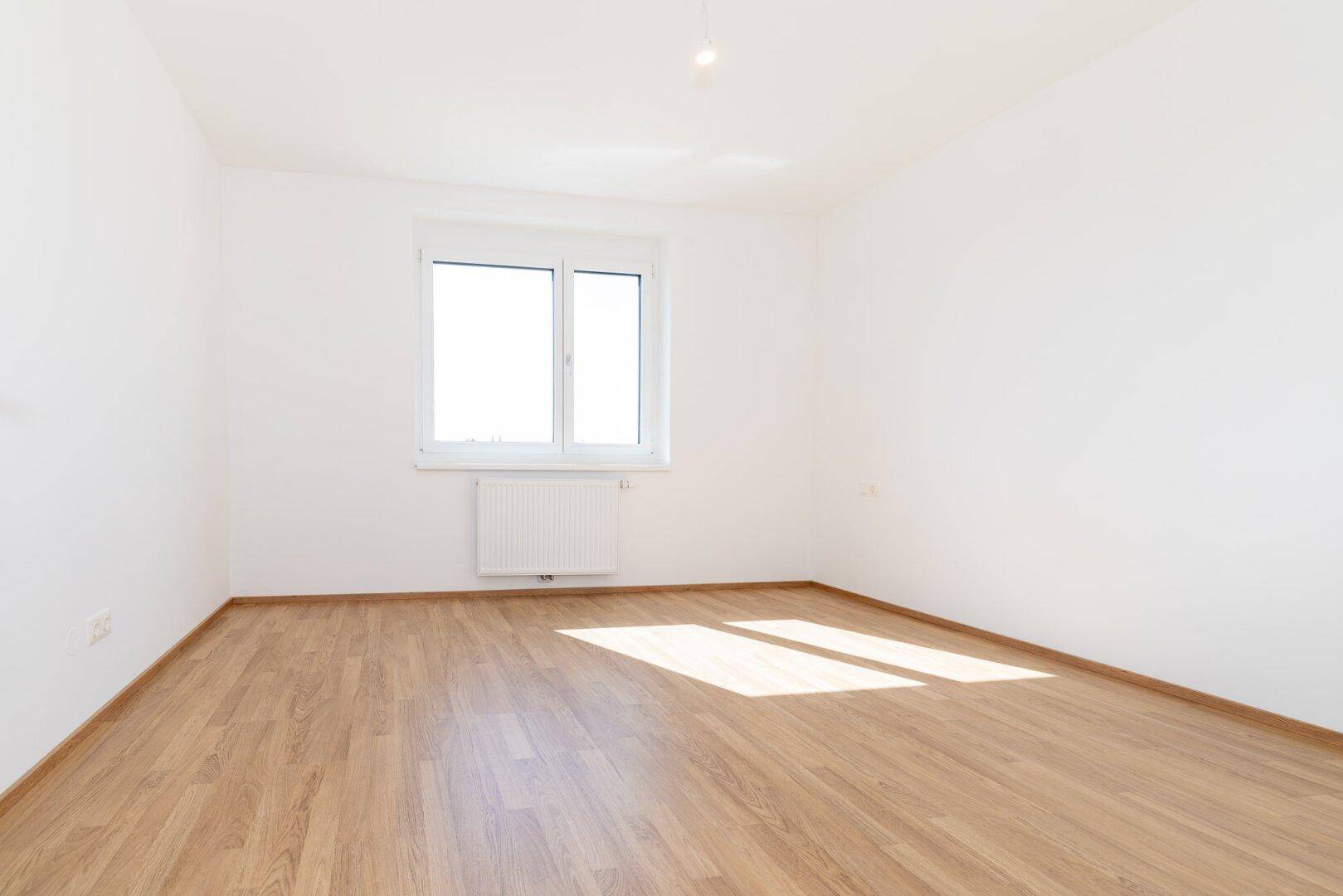 Helles Schlafzimmer mit Laminatboden und großem Fenster, das viel Tageslicht hereinlässt.