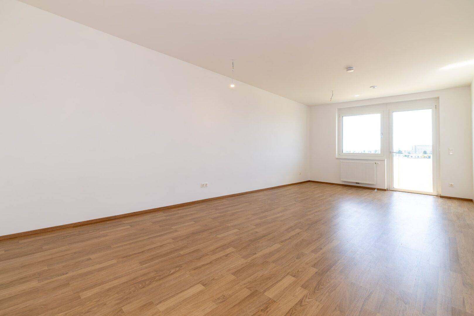 Helles, leeres Zimmer mit Laminatboden und Zugang zu einem Balkon.