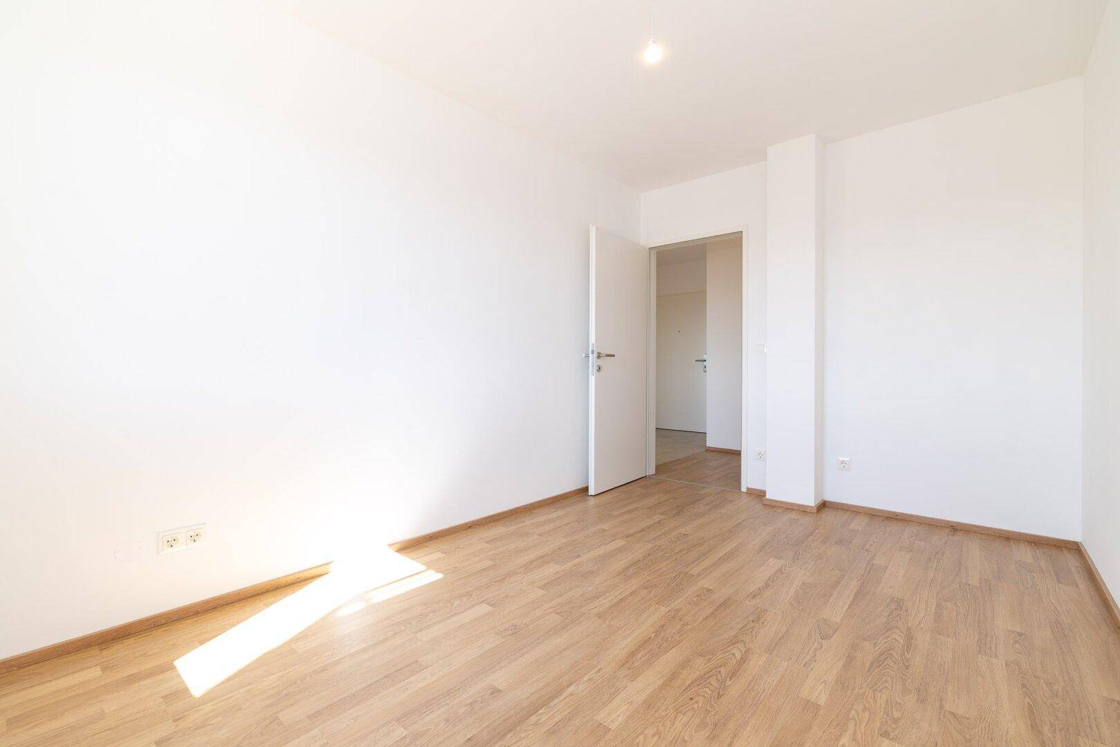 Helles Schlafzimmer mit Laminatboden und offener Tür, die den Blick in den Flur freigibt.