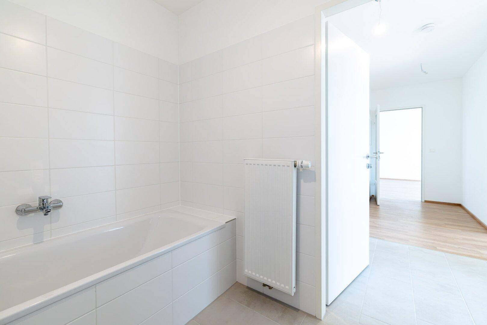 Helles Badezimmer mit Badewanne, weißen Fliesen und Heizkörper, mit Zugang zu einem weiteren Raum.