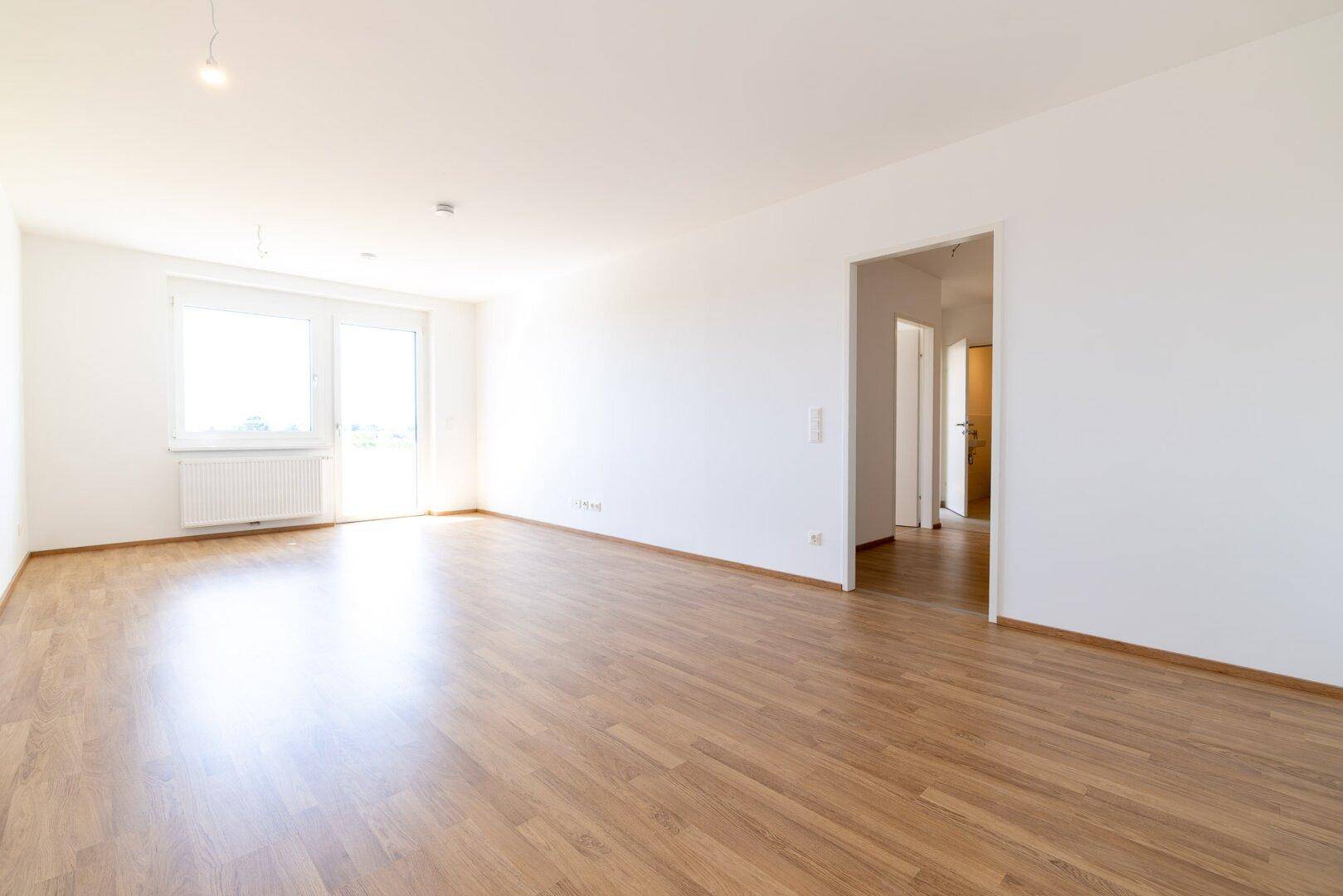 Geräumiges, leeres Wohnzimmer mit hellem Laminatboden und Zugang zu einem Balkon.