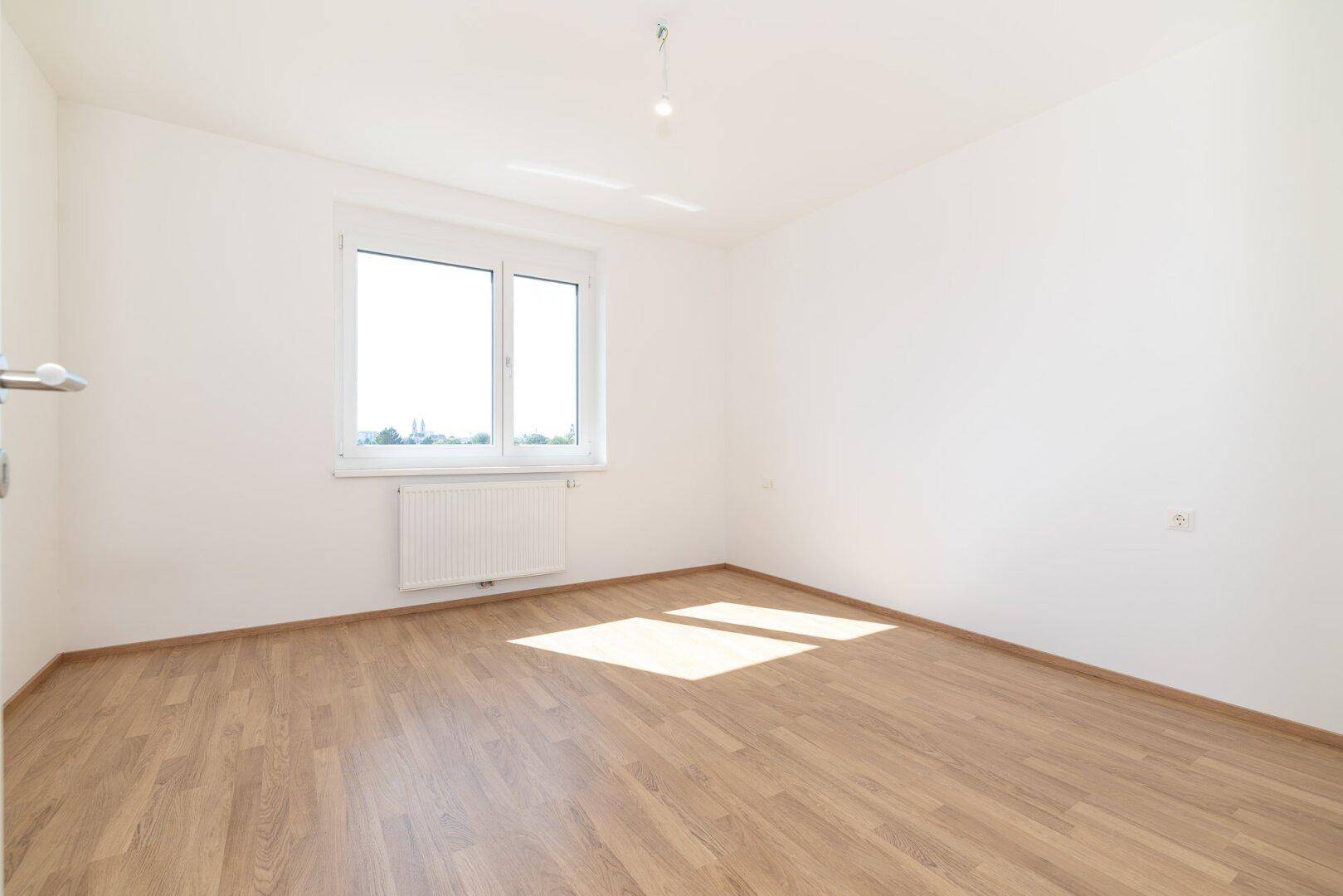 Helles Schlafzimmer mit Laminatboden und großem Fenster, das viel Tageslicht hereinlässt.