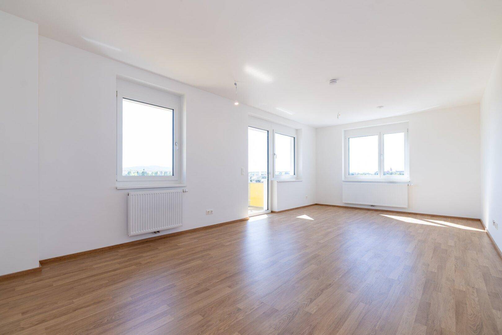 Geräumiges, helles Wohnzimmer mit großen Fenstern, Laminatboden und Zugang zum Balkon.