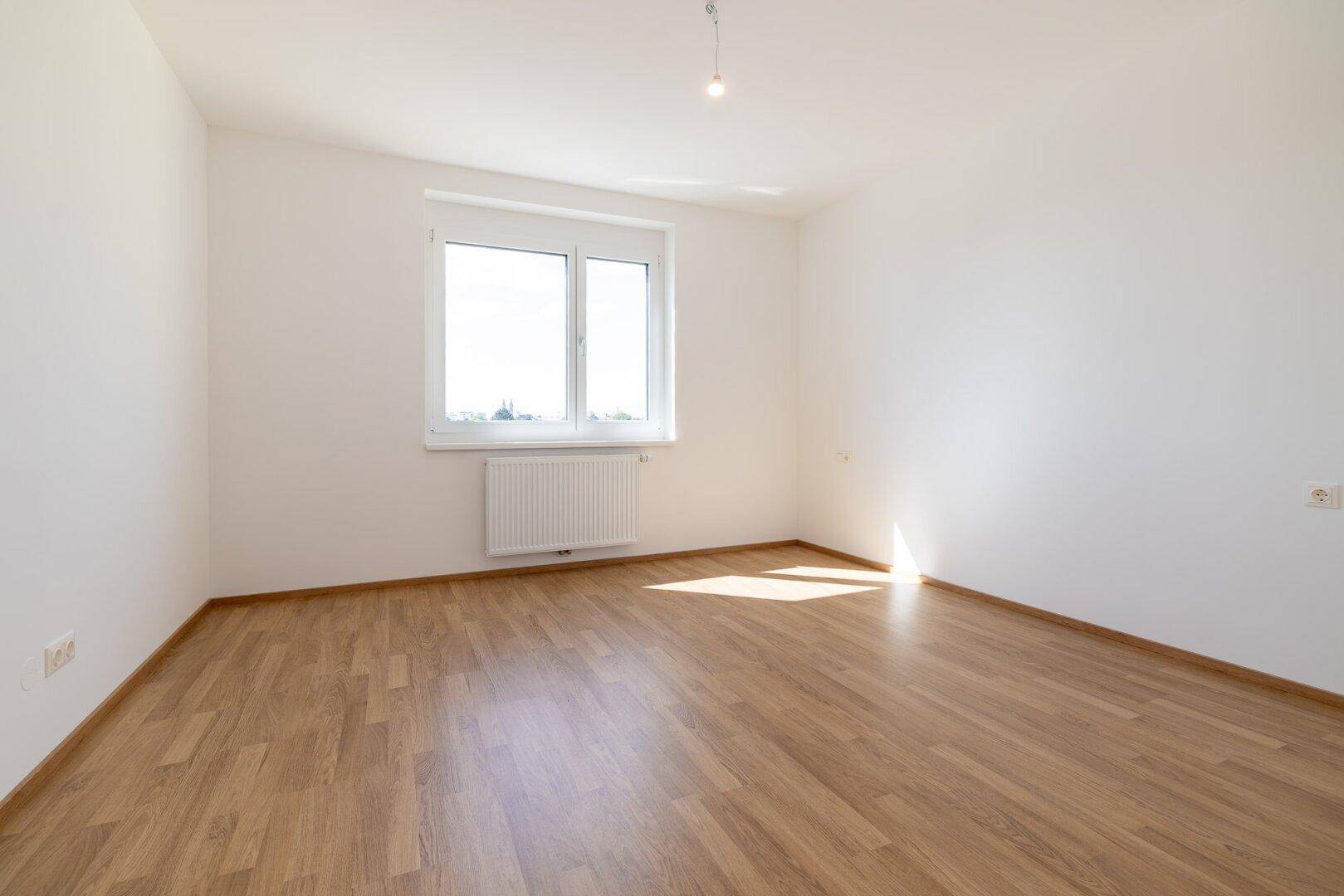 Helles Schlafzimmer mit Laminatboden, weiß gestrichenen Wänden und einem großen Fenster.