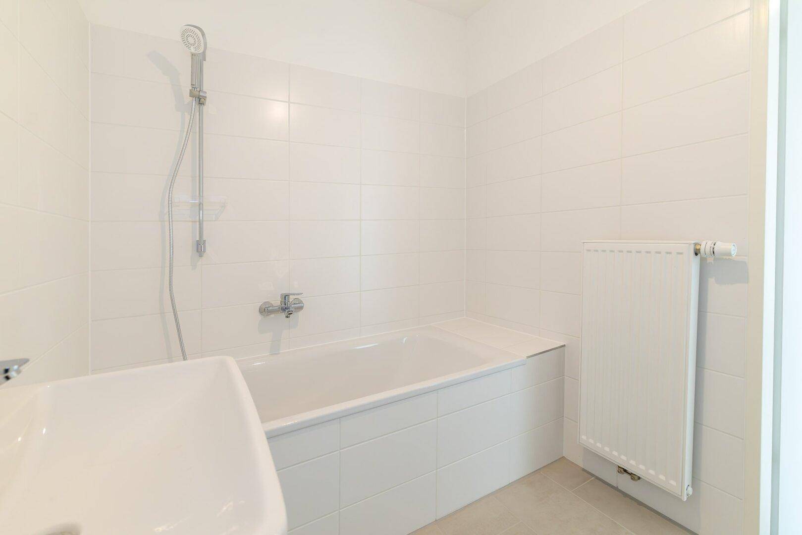 Helles Badezimmer mit weißen Fliesen, Badewanne, Waschbecken und Heizkörper.