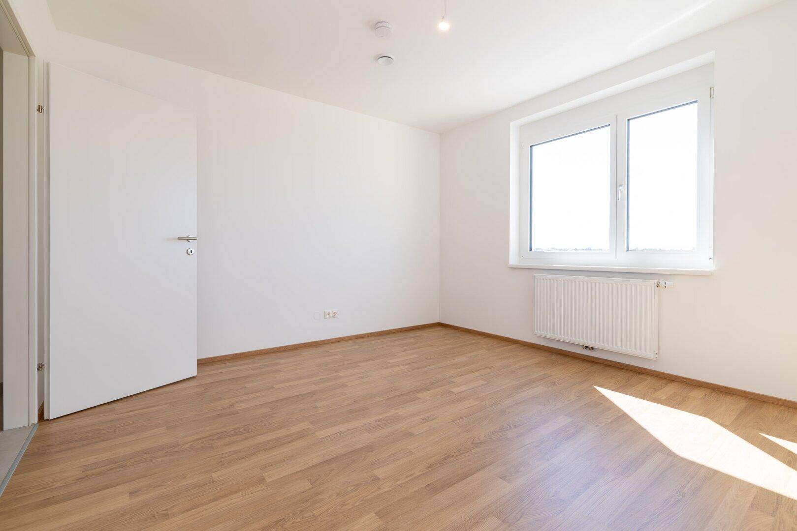 Geräumiges, helles Schlafzimmer mit Laminatboden, weiß gestrichenen Wänden und einer Tür.