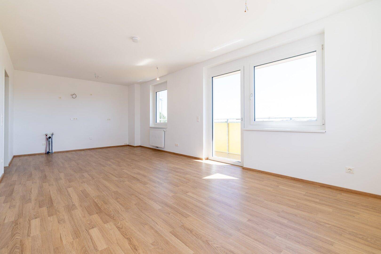 Heller Wohnbereich mit Laminatboden, großen Fenstern und Vorinstallationen für eine Küche.