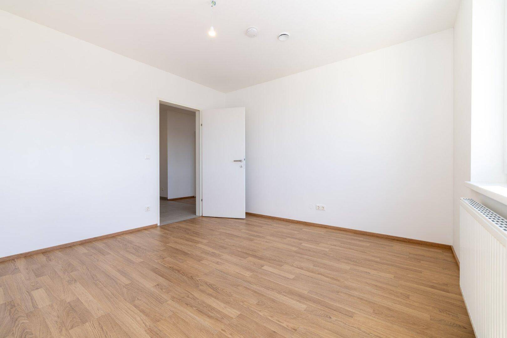 Helles Schlafzimmer mit Laminatboden, weiß gestrichenen Wänden und offenem Zugang zu einem weiteren Raum.