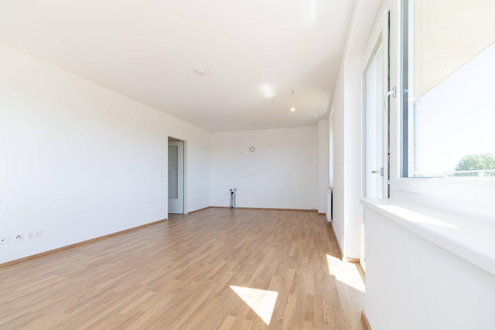 Heller Wohnbereich mit Laminatboden, großen Fenstern und Vorinstallationen für eine Küche.