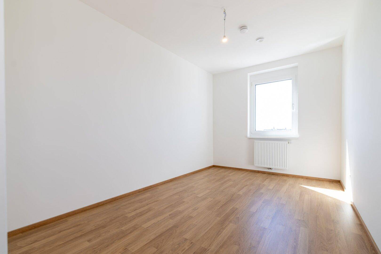 Helles Schlafzimmer mit Laminatboden, weiß gestrichenen Wänden und einem Fenster.