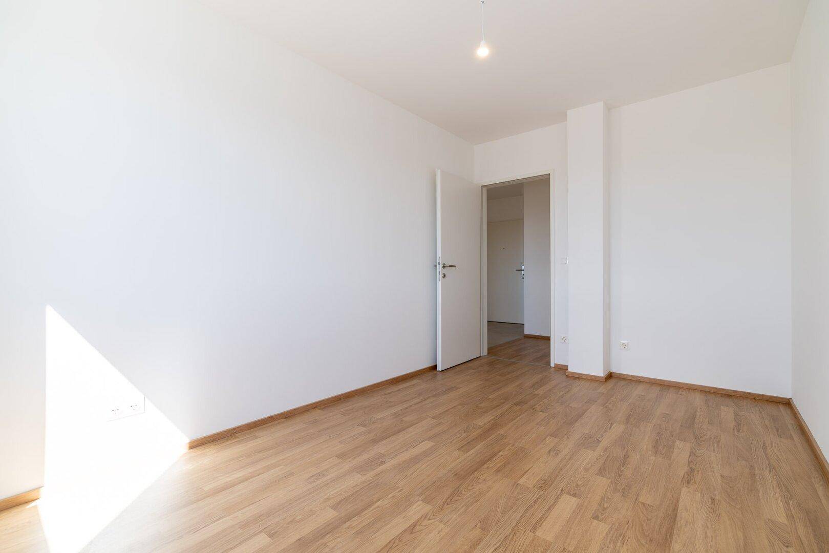 Helles Schlafzimmer mit Laminatboden, weiß gestrichenen Wänden und offenem Zugang zu einem weiteren Raum.