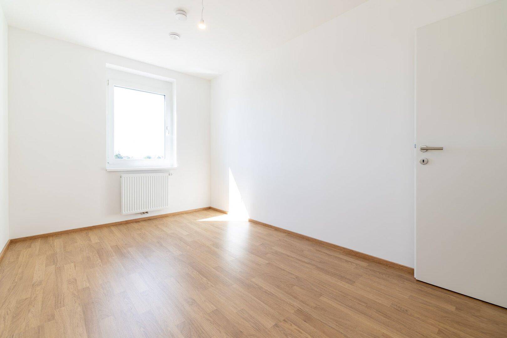 Geräumiges, helles Schlafzimmer mit Laminatboden, weiß gestrichenen Wänden und einer Tür.