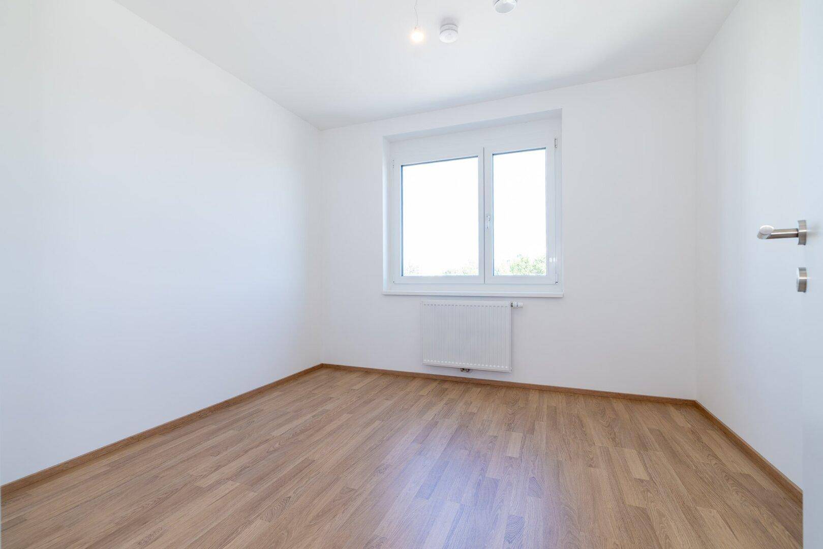 Helles Schlafzimmer mit Laminatboden, weißen Wänden und großem Fenster für viel Tageslicht.