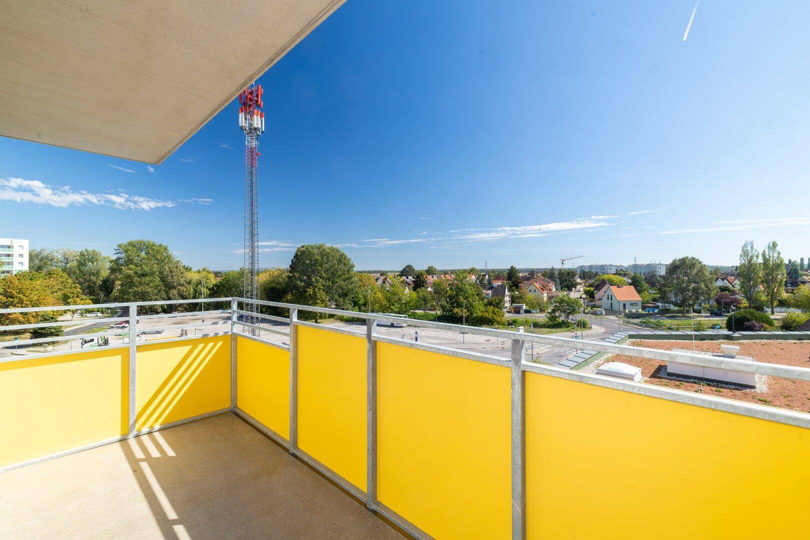 Sonniger Balkon mit gelben Brüstungselementen und Blick auf die Umgebung und einen Mobilfunkmast.