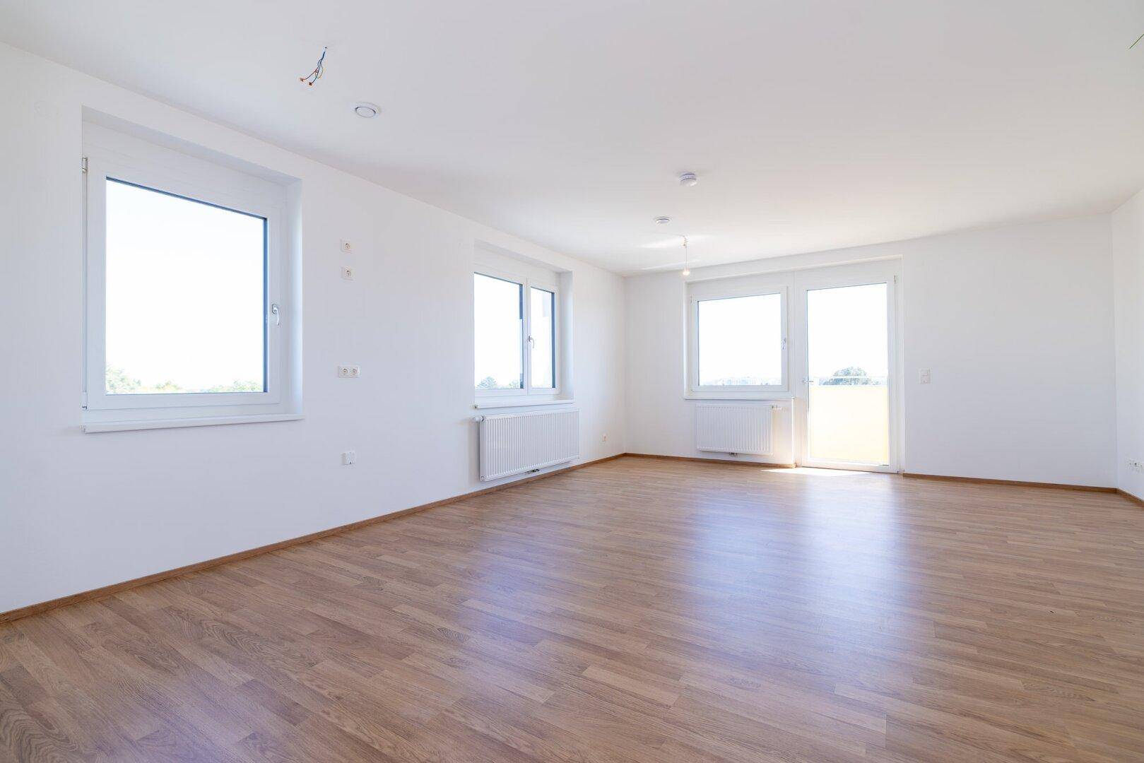 Geräumiges, helles Wohnzimmer mit großen Fenstern, Laminatboden und Zugang zum Balkon.