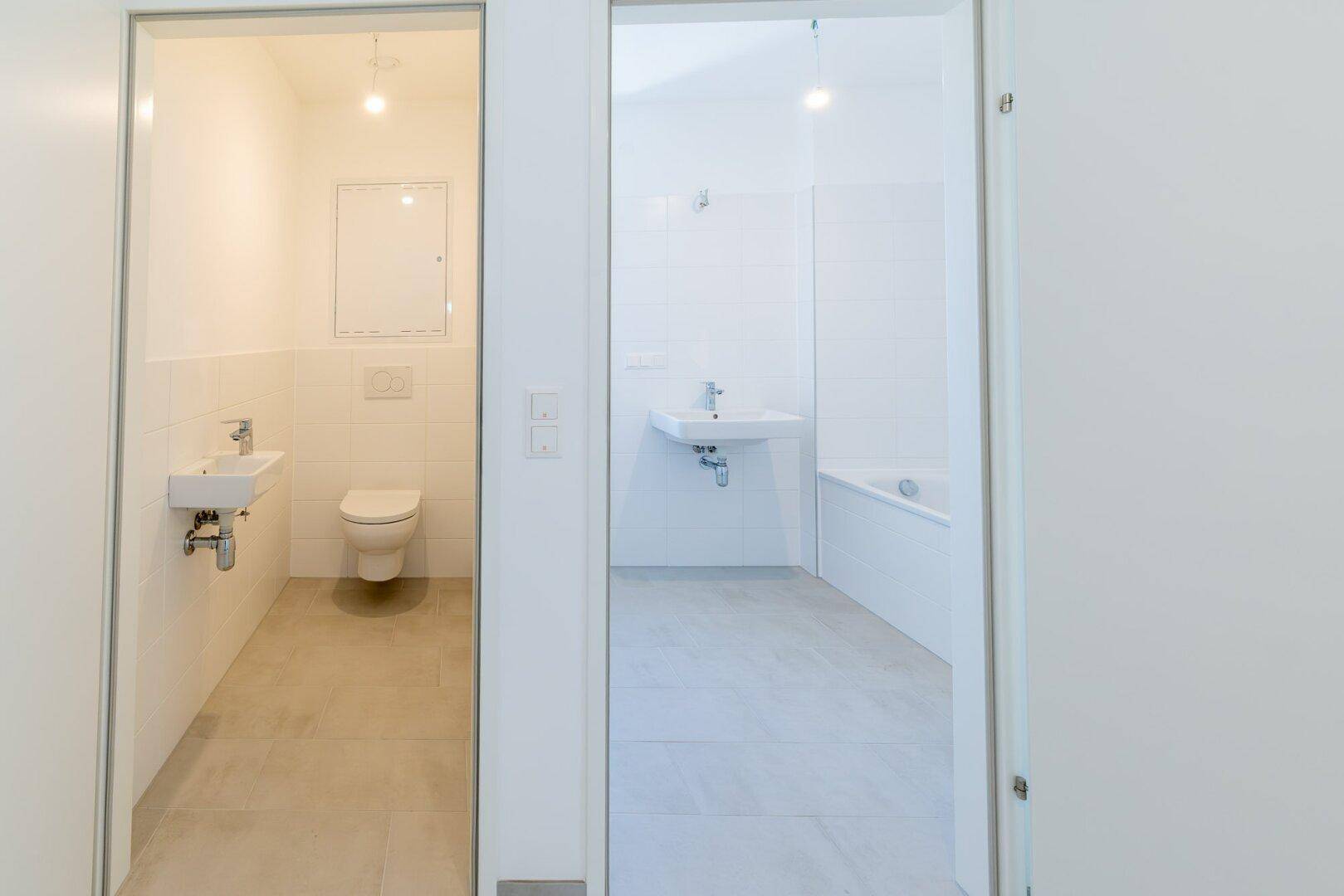 Blick in ein modernes Gäste-WC und ein Badezimmer mit weißen Fliesen und Sanitärobjekten.