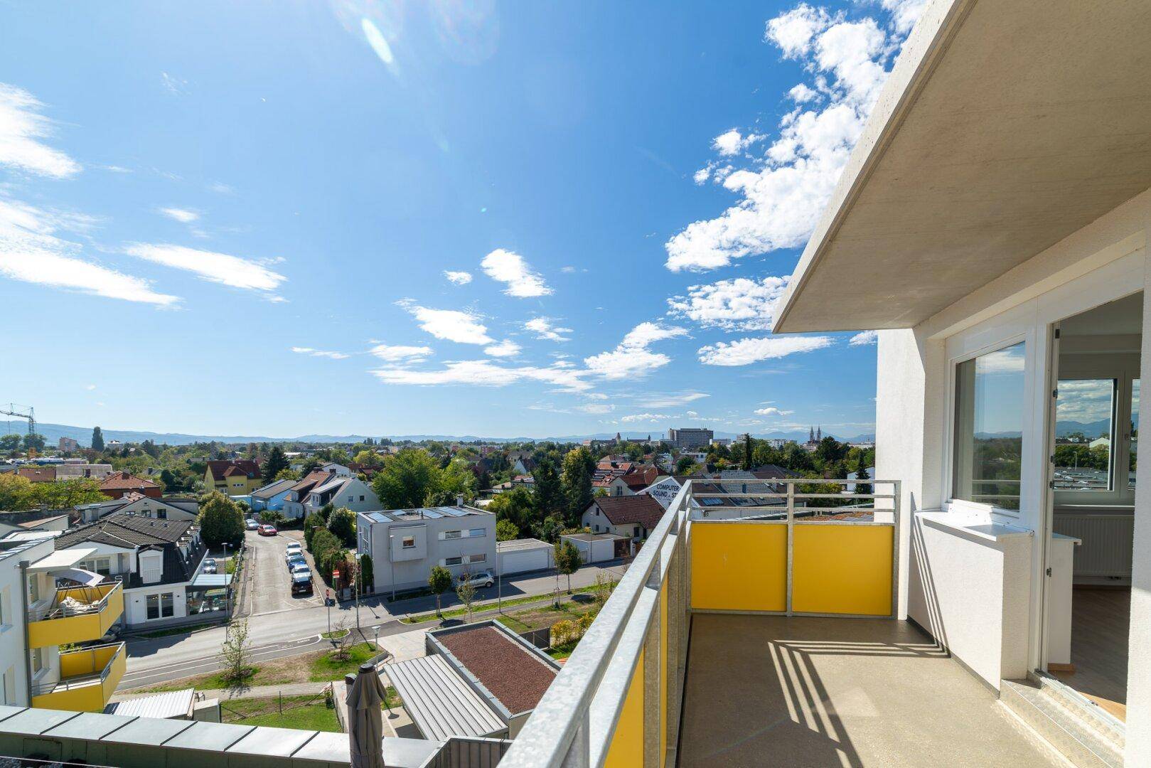 Sonniger Balkon mit gelbem Geländer und weitem Blick über die Wohnsiedlung und die Straße.