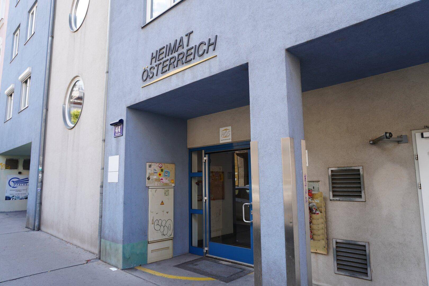 Eingangsbereich eines Gebäudes mit der Aufschrift "HEIMAT ÖSTERREICH", blauer Glastür und Überwachungskameras.