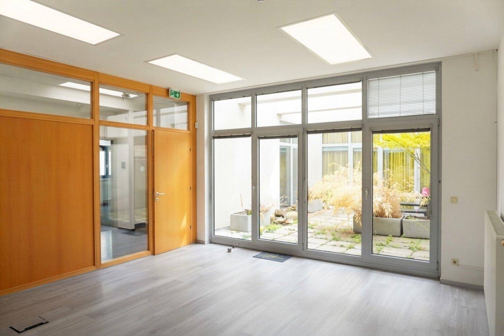 Geräumiges Büro mit hellem Laminatboden und großen Fenstern mit Blick auf einen begrünten Innenhof.