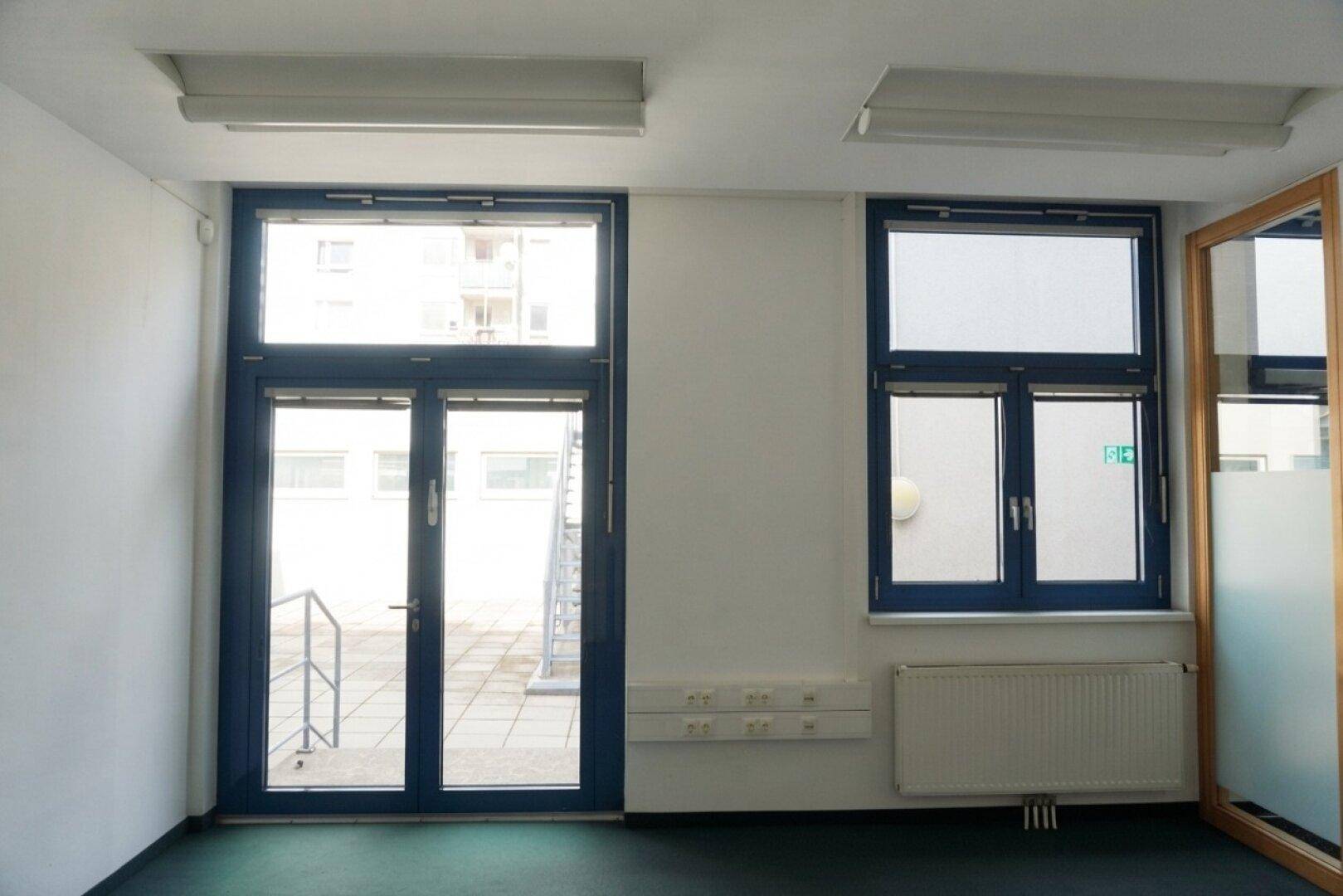 Heller Büroraum mit dunklem Teppichboden, Heizkörper und blauen Fenstern mit Zugang zur Terrasse.