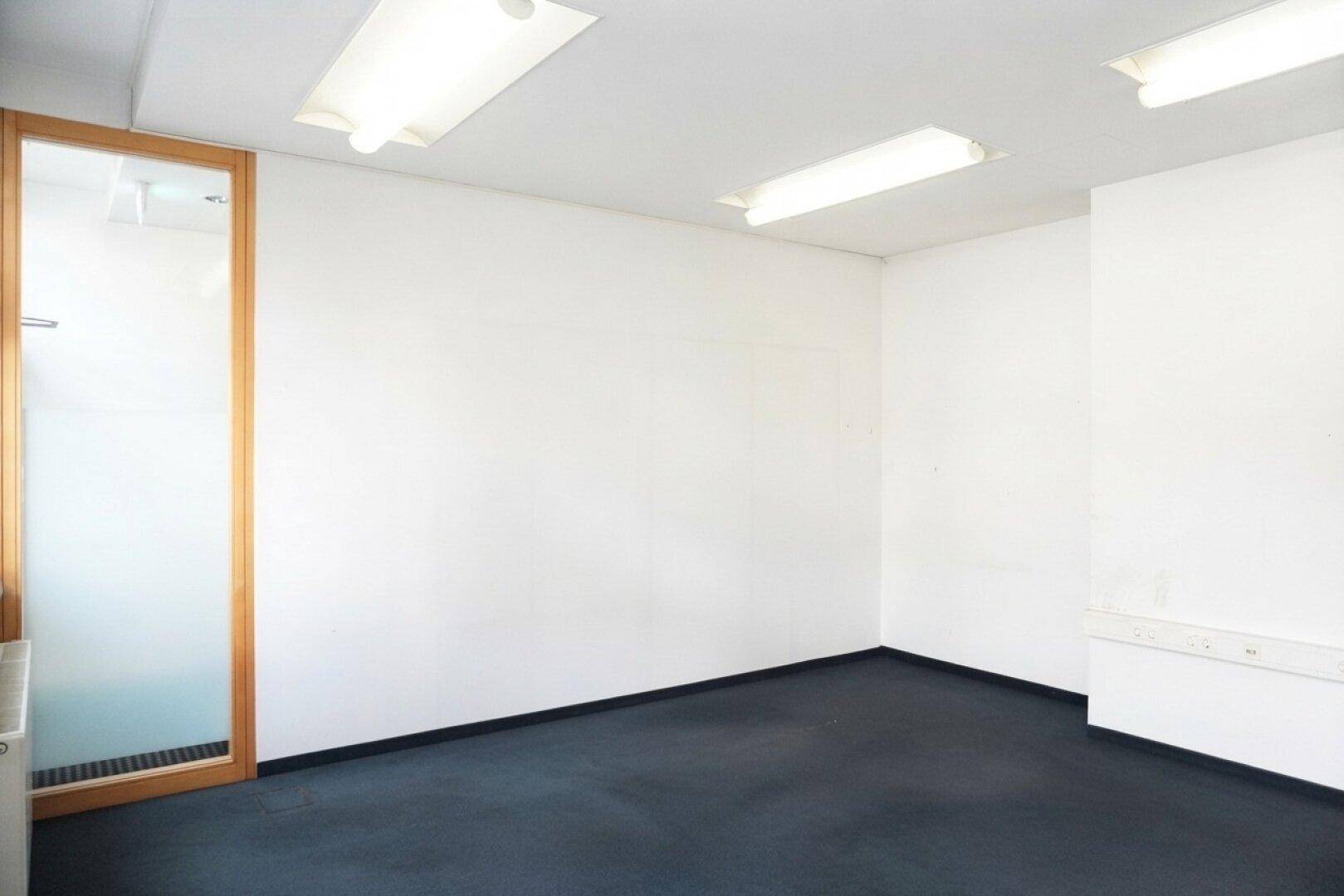Schlichter Büroraum mit dunklem Teppichboden und weißen Wänden, ausgestattet mit funktionalen Deckenleuchten.