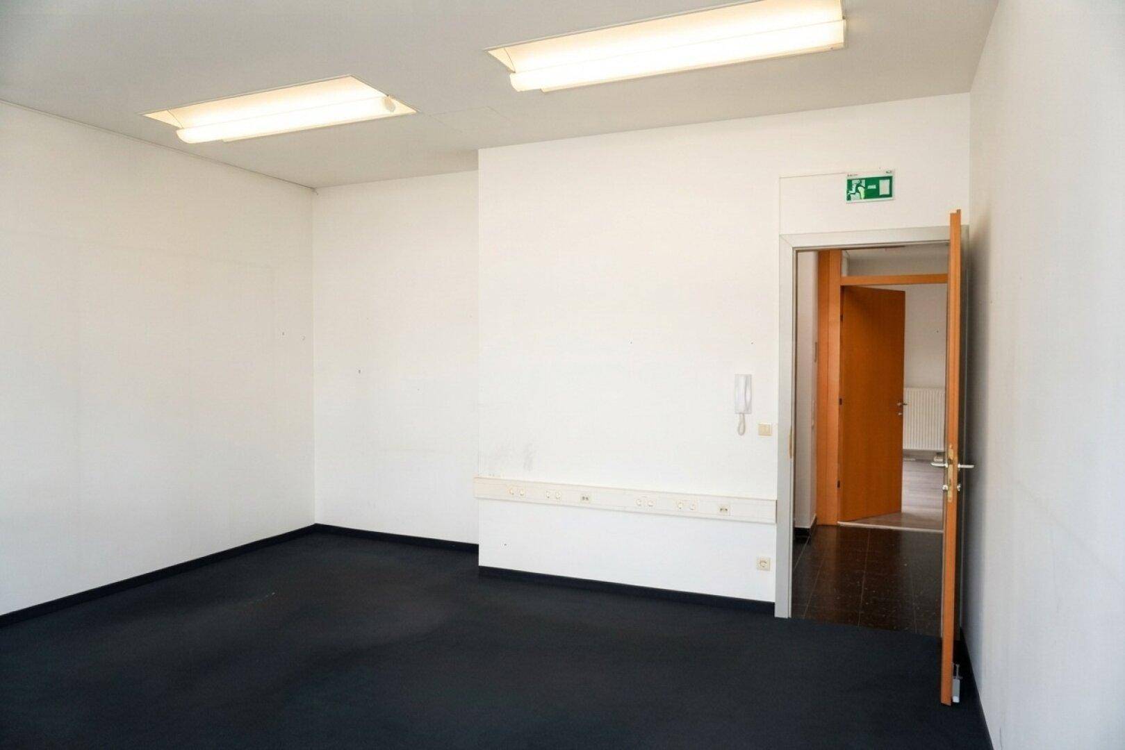 Leerer Büroraum mit dunklem Teppichboden und weiß gestrichenen Wänden, mit Tür zum angrenzenden Flur.